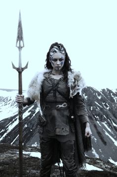 a woman with makeup on holding a large spear in front of snow covered hills and mountains