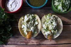 two fish tacos are sitting on a wooden table next to some dipping sauces