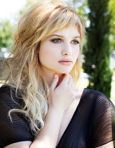 a woman with long blonde hair wearing a black top and posing for a photo in front of some trees