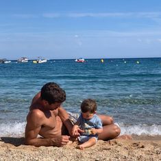 a man and child sitting on the sand at the beach with boats in the water