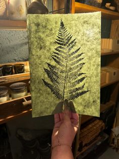 a hand holding up a piece of paper with a fern leaf on it in front of shelves