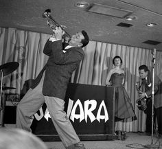 a man in a suit and tie playing trumpet on stage with other people behind him