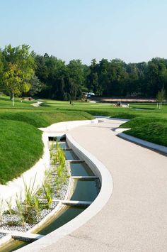 a curved walkway with water running through it in the middle of a grassy park area