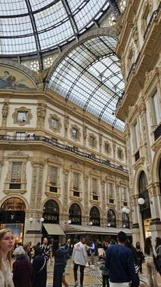 many people are walking around in the shopping mall with glass ceiling and high arched windows