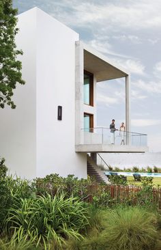 two people standing on the balcony of a modern house with grass and shrubs surrounding it