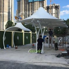 two men are standing under a white tent in the middle of a construction area with tall buildings behind them