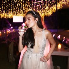 a woman in a white dress drinking from a wine glass at a party with lights hanging above her