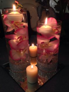 two clear vases with pink flowers and lit candles on a black table top in front of a white tea light