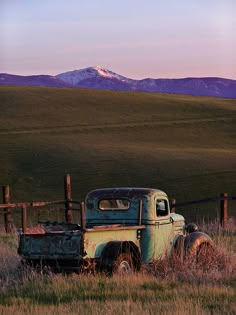 an old truck sitting in the middle of a grassy field with mountains in the background