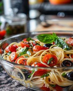 a glass bowl filled with pasta, tomatoes and olives on top of a counter