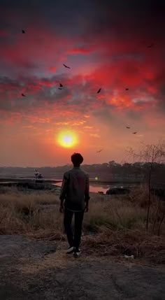 a man standing on top of a dirt field under a red and blue sky with birds flying in the air