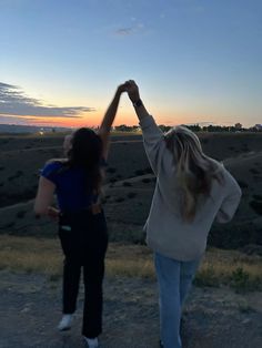 two women holding hands in the middle of a dirt road at sunset, with their arms raised