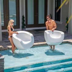 a man and woman standing in front of a swimming pool with two giant white chairs