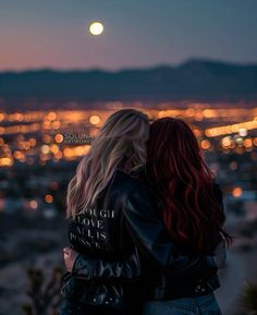 two women standing on top of a hill looking out at the city lights in the distance