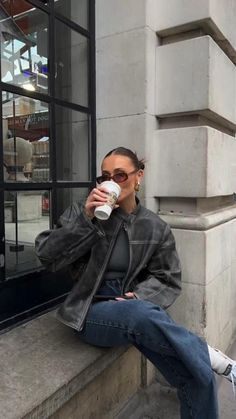 a woman sitting on the curb drinking from a coffee cup while wearing sunglasses and a leather jacket