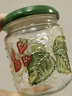 a hand holding a glass jar with strawberries and leaves painted on the side,