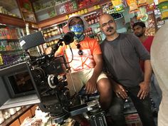 two men sitting in front of a camera on a tripod with food behind them