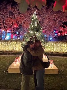 two people standing in front of a christmas tree with lights on the trees behind them