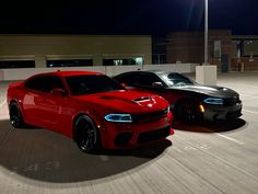 two red and black cars are parked in an empty parking lot at night with the lights on