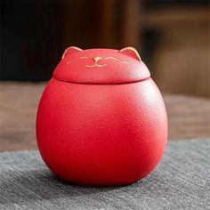 a red cat shaped container sitting on top of a gray cloth covered table with a wooden chair in the background