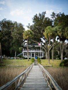 a wooden walkway leading to a large white house in the distance with tall grass and trees