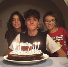 three people sitting in front of a cake with lit candles on it and one person standing next to the cake