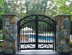 an iron gate is open to a swimming pool in the back yard, surrounded by stone walls and trees