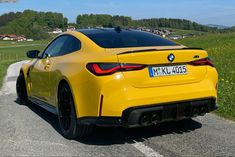 the rear end of a yellow bmw z4 driving down a country road with houses in the background