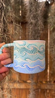 a hand holding a blue and white coffee mug in front of some branches with moss