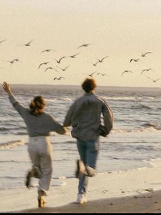 two people running on the beach with seagulls flying in the sky behind them