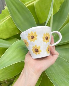 a hand holding a coffee cup with yellow flowers on it and green leaves in the background