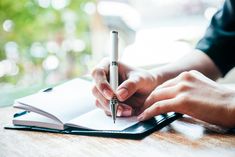 a person writing on a notebook with a pen in their hand and an open book behind them