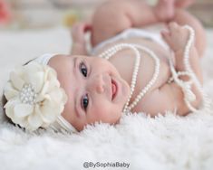 a baby laying on top of a white rug wearing pearls and a flower in her hair