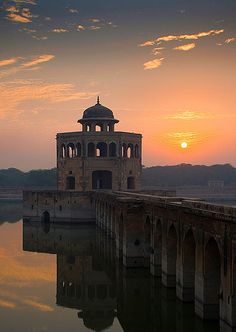 the sun is setting behind an old building on the water's edge in front of mountains