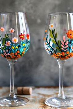 Two wine glasses with floral patterns painted on them, sitting on a wooden surface with a cork nearby. Diy Glassware Projects, How To Paint On Wine Glasses, Wine Glass Design Ideas, Painting Ideas On Glass Cups, Acrylic Wine Glass Painting, Art And Wine Painting Ideas, Wine Glass Ideas Diy, Hand Painted Glasses Diy, Painting On Wine Glasses Diy