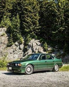 a green car parked on the side of a dirt road next to some tall trees