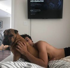 a person laying in bed with a dog on his chest and a television mounted to the wall behind them