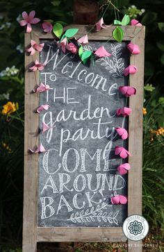 a chalkboard sign with pink and green paper flowers on it that says welcome to the garden party come around back