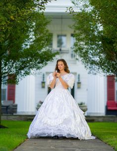 a woman in a white wedding dress standing on a sidewalk with trees and grass behind her