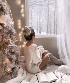 a woman sitting on the floor next to a christmas tree reading a book and drinking coffee