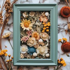 an arrangement of flowers in a frame on a marble surface with dried grass and sunflowers