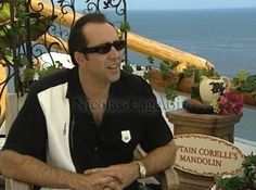 a man wearing sunglasses sitting on top of a chair next to a sign that says captain corelli's mandolin