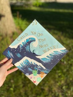 a hand holding up a blue graduation cap that says catch ya on the next wave