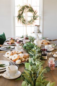 the table is set for christmas dinner with plates and cups on it, along with greenery