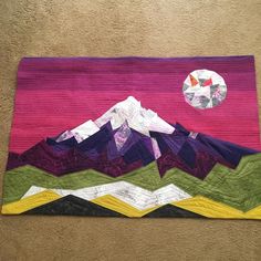 a quilted wall hanging with mountains and a moon in the sky on it's side