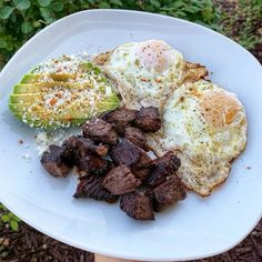 an egg, steak and avocado on a white plate in the middle of some grass