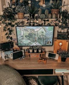 there is a computer screen on the desk with many plants and other things around it