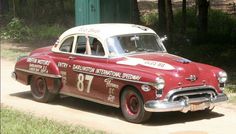 an old red and white car driving down a dirt road