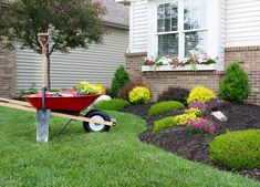 a red wheelbarrow in front of a house with flowers and shrubs around it