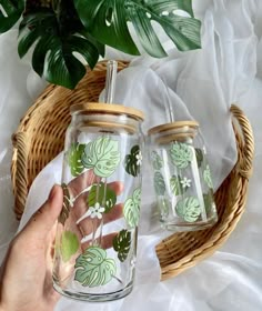 a hand holding a jar with green leaves on it next to a wicker basket